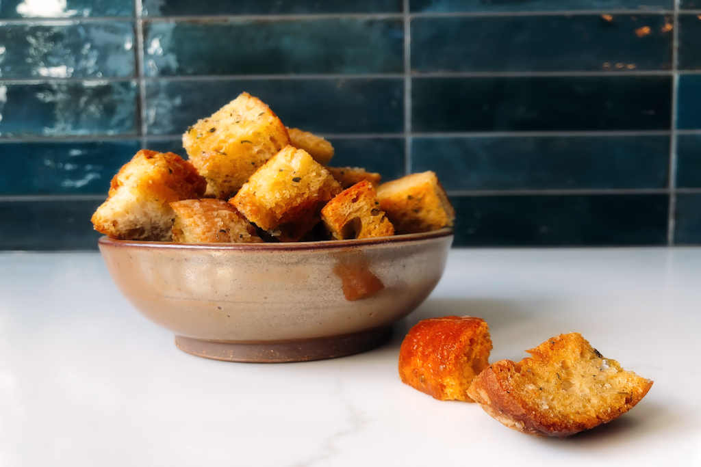 Bowl of croutons on white counter with blue tile background.